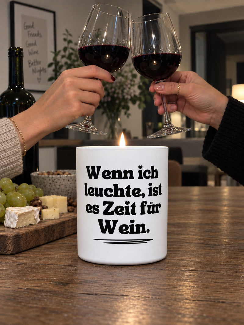 Zwei Freundinnen stoßen mit Rotwein an, davor die Duftkerze mit dem Spruch Wenn ich leuchte, ist es Zeit für Wein
